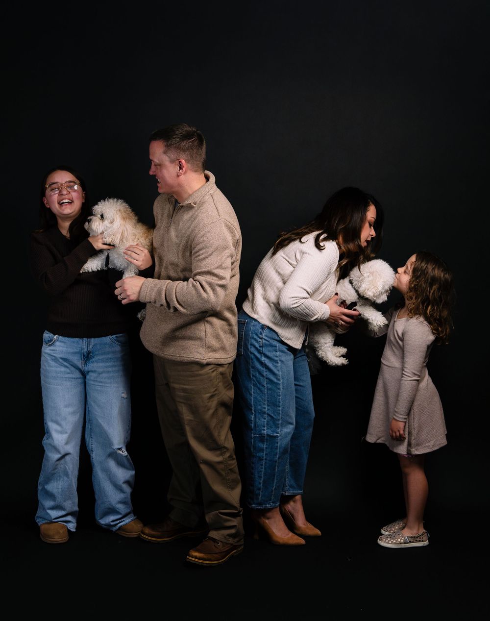 Family portrait with two adults, a child, and two white fluffy dogs posing playfully against a dark studio background.