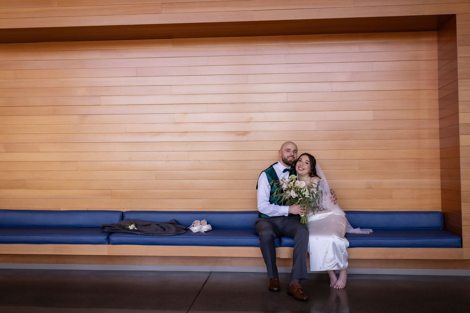 Bride and groom sitting together on a blue bench holding a bouquet at their wedding.