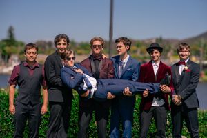 Group of seven young men in formal attire posing outdoors, one being carried horizontally, before a prom. San Diego, CA