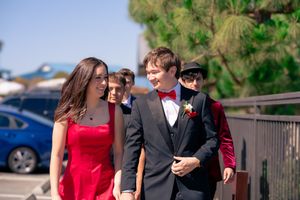 Teens dressed in formal prom attire walk together outdoors, boy in suit with red bow tie, girl in red dress. San Diego, CA