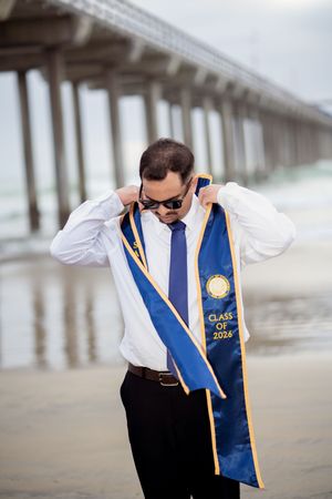 Man in white shirt and blue tie wearing Class of 2026 graduation sash on beach near pier. La Jolla, CA
