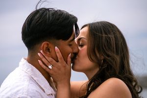 Couple sharing a romantic kiss outdoors, woman wearing an engagement ring, against a cloudy sky backdrop. Carlsbad, CA