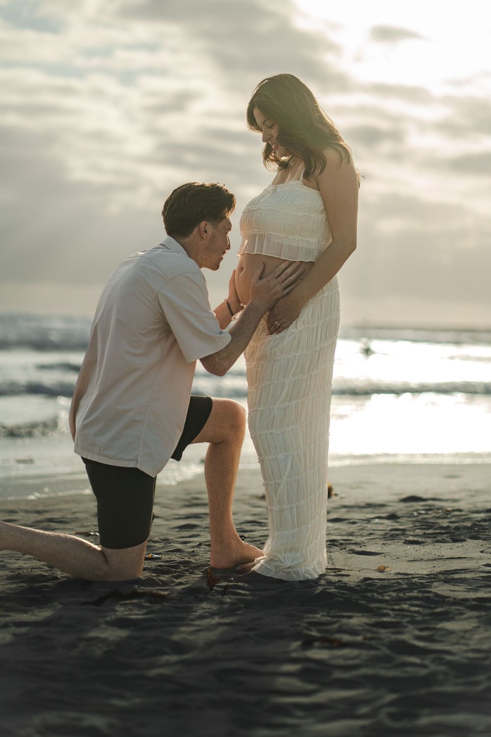 A man kneels on a black sand beach touching a pregnant woman's belly at sunset. Oceanside, CA