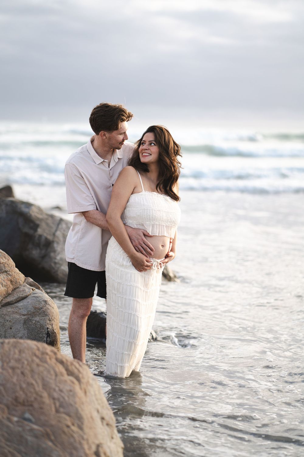 Pregnant couple standing in ocean waves at beach, man embracing woman in white dress while holding her baby bump. Oceanside, CA