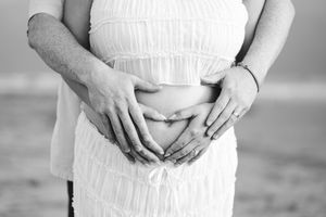 Black and white photo of pregnant woman in white dress with partner forming heart shape on baby bump at beach. Oceanside, CA
