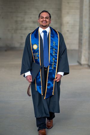 UC San Diego graduate smiling in black gown and blue satin stole walking confidently during graduation photos. La Jolla, CA