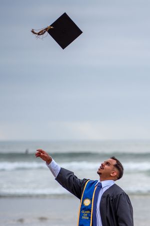 Graduate in cap and gown tosses grad cap into sky on beach, wearing Class of 2026 stole. La Jolla, CA