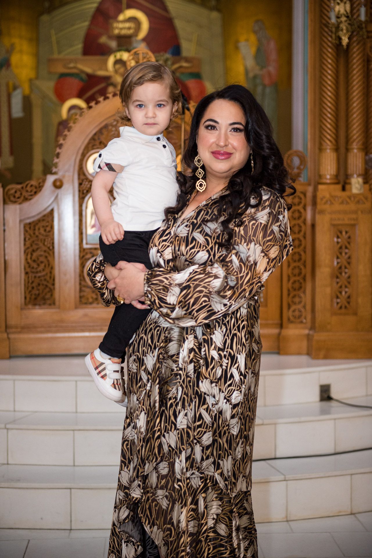 Woman in animal print dress holds young child in front of church iconostasis.