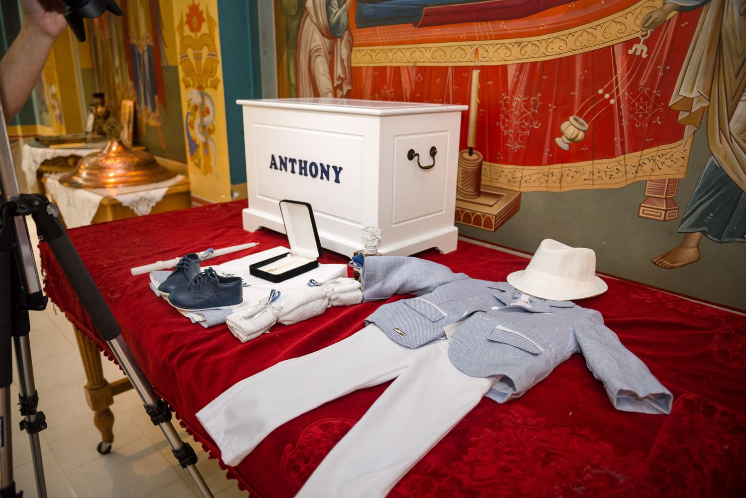 White personalized box labeled Anthony with baptism outfit and accessories on red velvet.