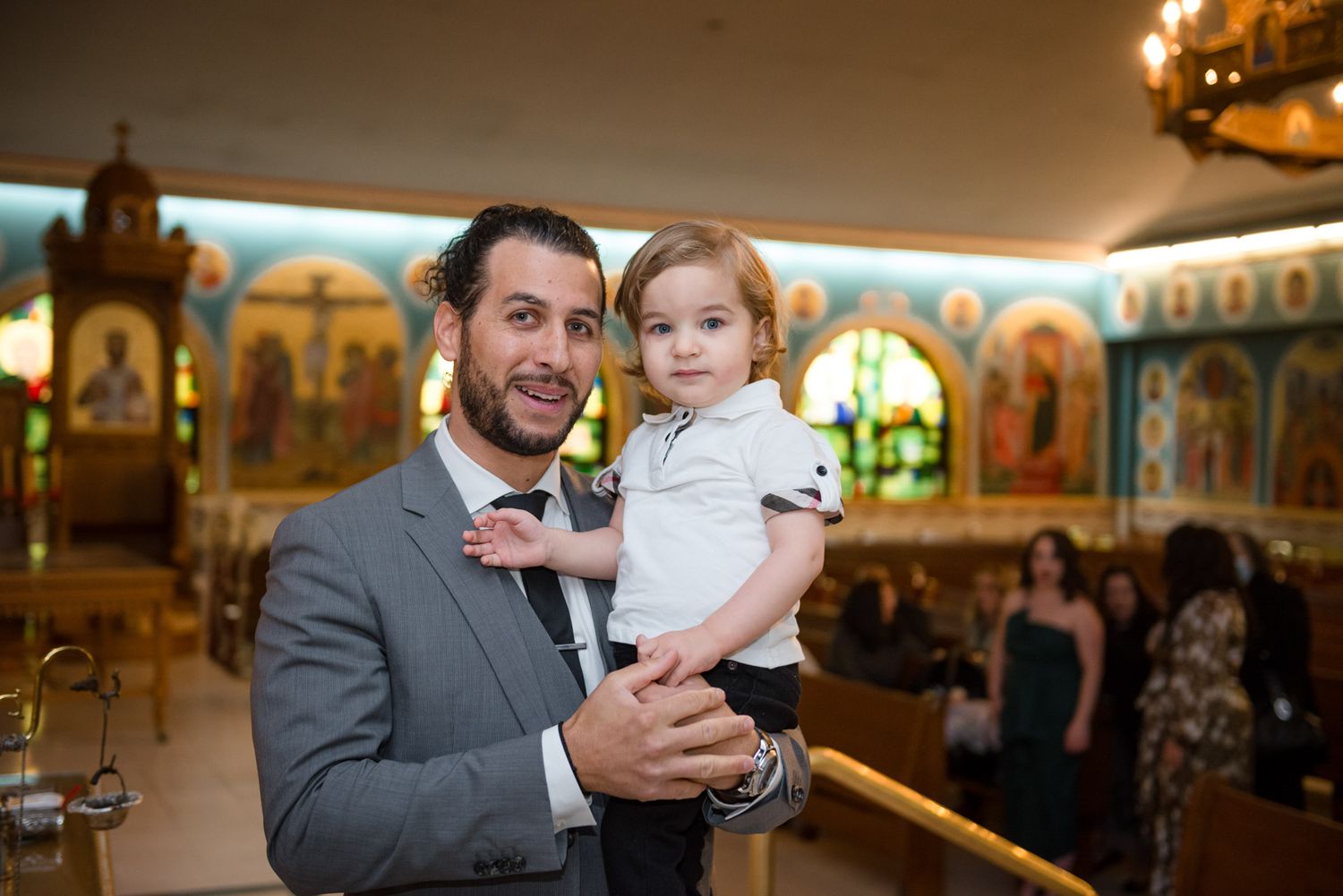 Father in gray suit holds young child inside decorated Orthodox Christian church.
