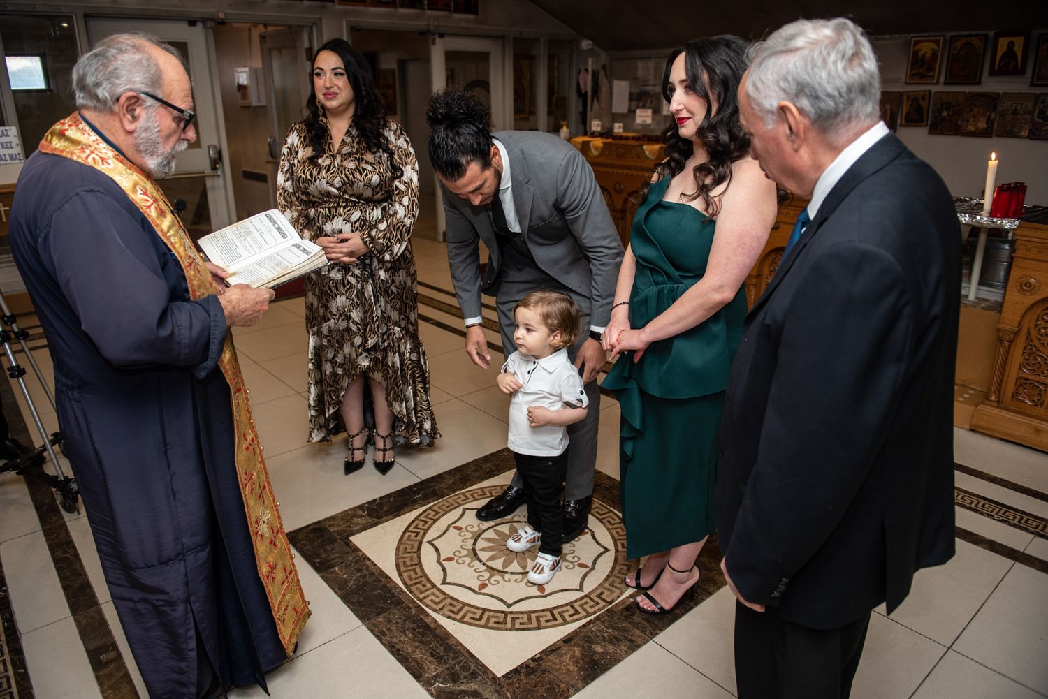 Family gathered around a young toddler during an Orthodox Christian baptism ceremony with a priest.