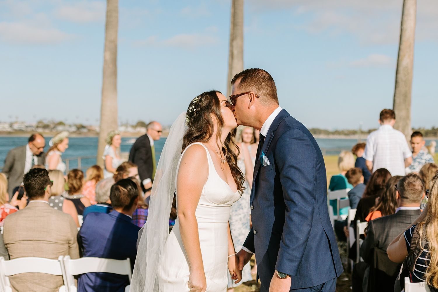 Bride and groom share first kiss at outdoor waterfront wedding ceremony with guests seated in white chairs at Tower Beach Club.