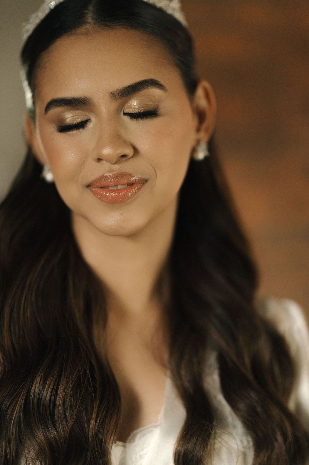 A bride with glittery eye makeup, glossy lips, pearl earrings, and a white headpiece poses with eyes closed.
