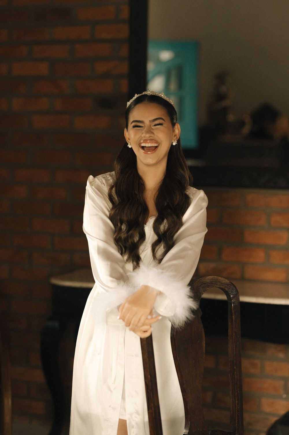 A smiling woman in a white satin robe with feather cuffs and a headband poses joyfully in a warmly lit room.