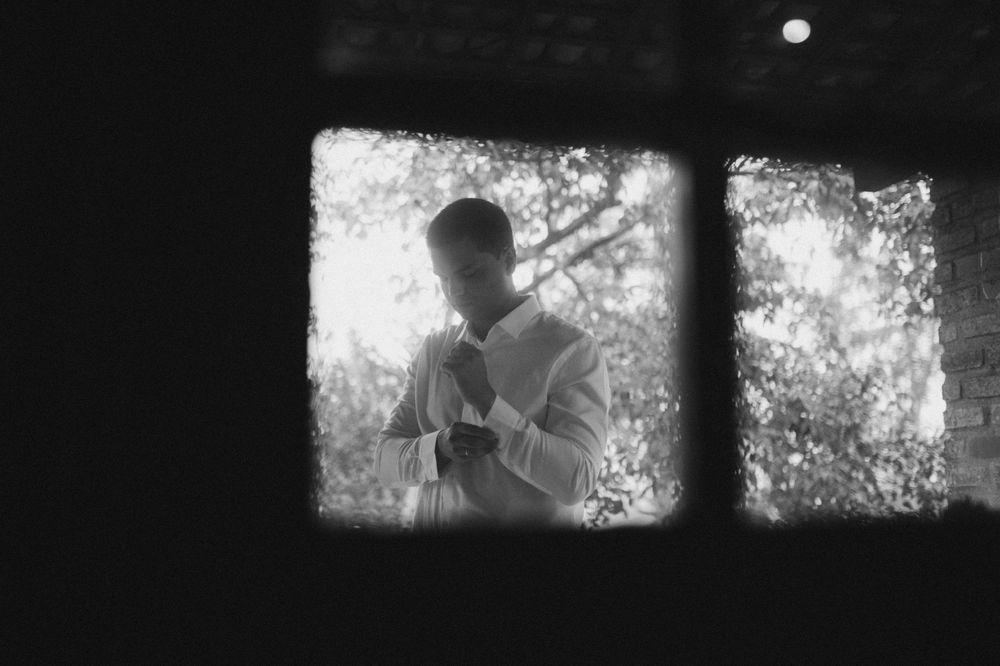 Black and white photo of a man adjusting his shirt, framed by a dark window with trees in background.