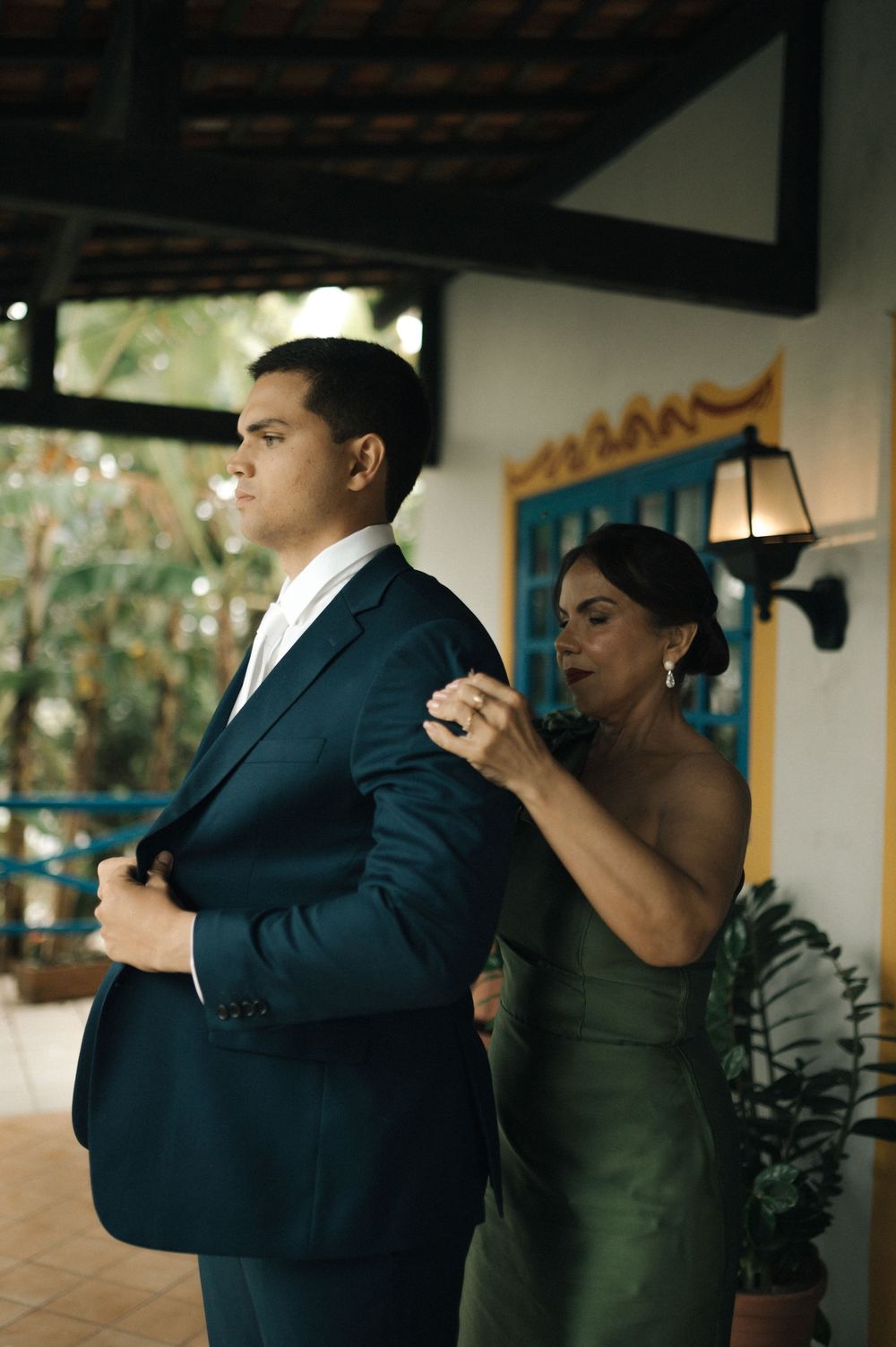 A woman in a green dress helps a man in a navy suit adjust his jacket at an elegant outdoor venue.