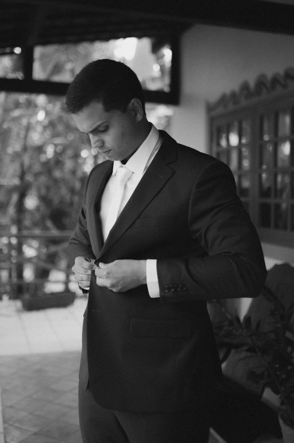 Groom in black suit and white tie buttoning jacket while looking down in elegant outdoor venue.