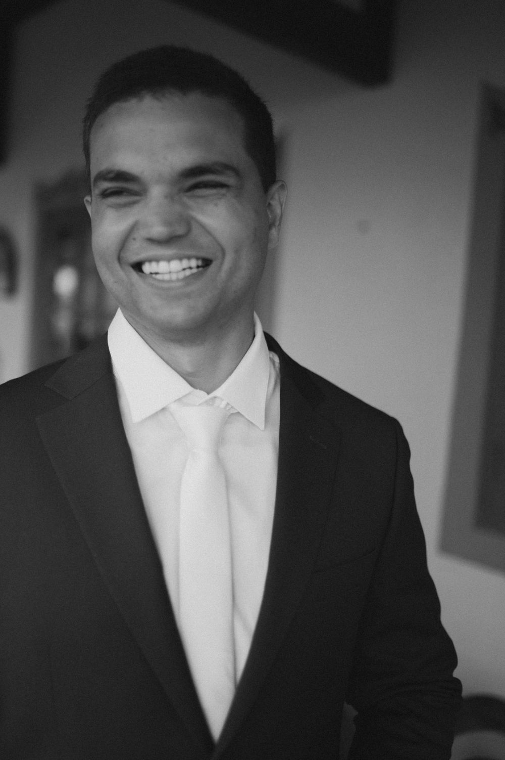 Smiling groom in black suit and white tie posing for portrait photo at wedding venue.