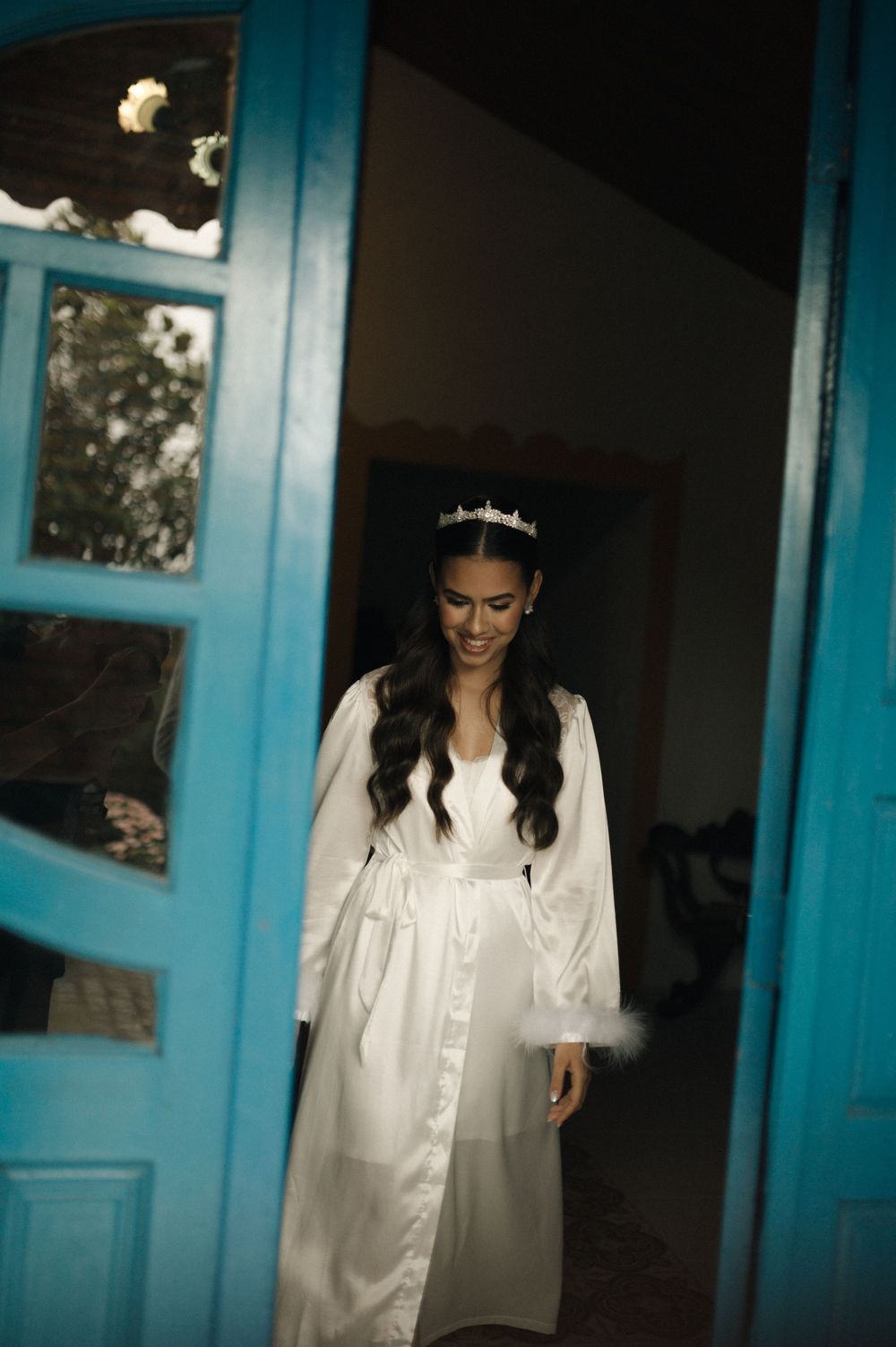 Bride in white robe and tiara smiling by blue door.