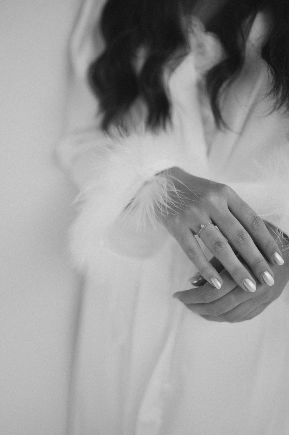 Black and white photo of a bride in a white feather-trimmed robe, showcasing her manicured hands and engagement ring.