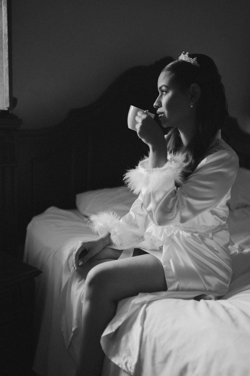 Black and white photo of a bride in silk robe and tiara sipping from a cup while sitting on a bed.