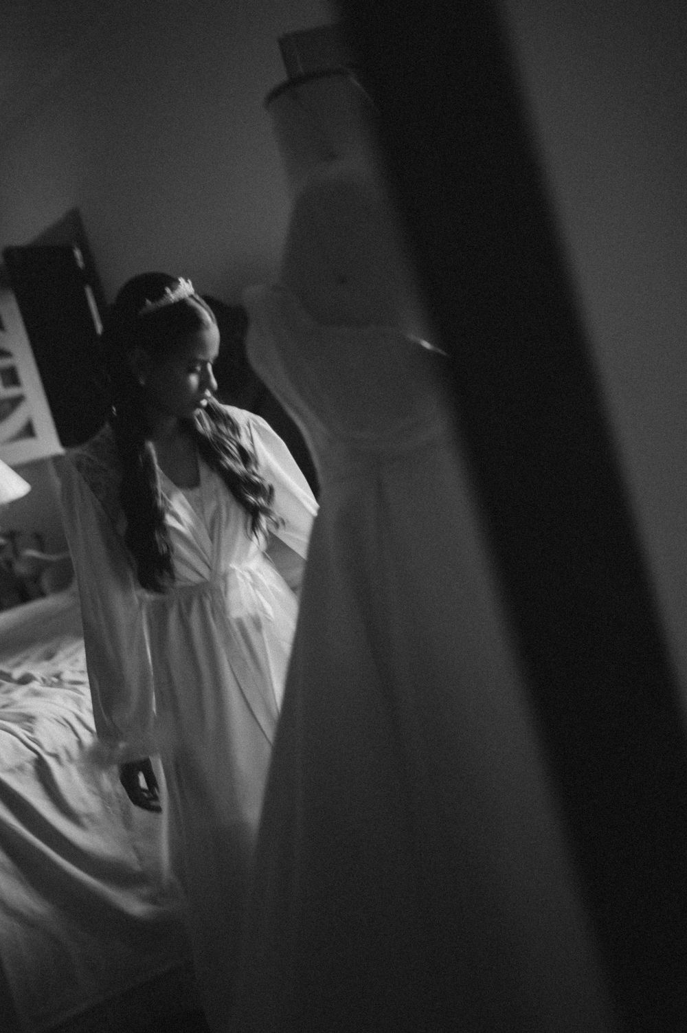 Black and white photo of a bride in a white gown and tiara standing in a dimly lit bedroom with her wedding dress hanging nearby.