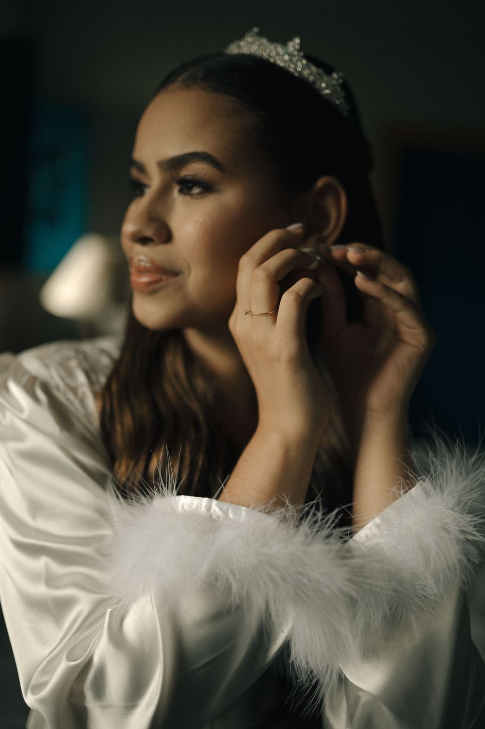 Bride in white satin robe with feather trim adjusts earring, wearing crystal tiara, in elegant pre-wedding preparation.