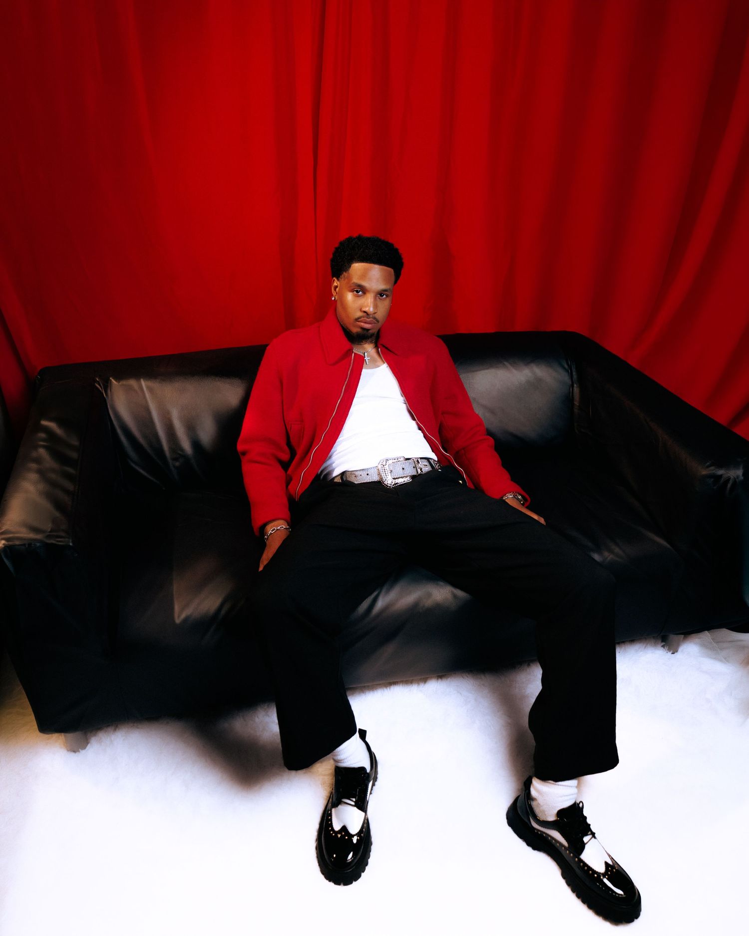 A young man in a red jacket poses confidently against a red curtain backdrop, wearing a watch and chain accessories.