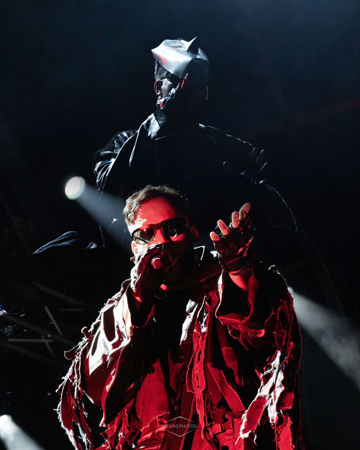 Performer in dramatic red shredded coat gestures on stage under spotlights during a live concert performance.