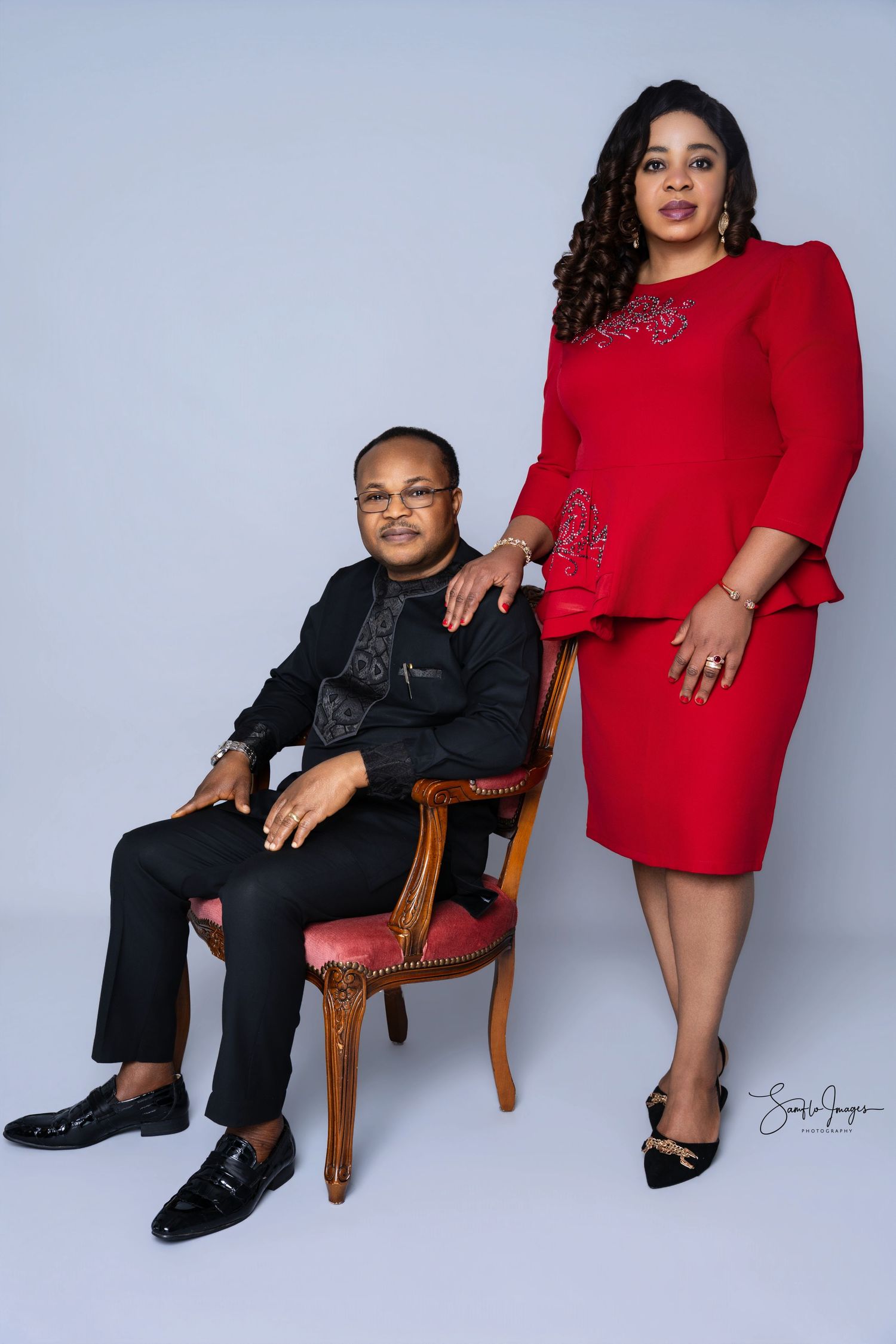 A man in black sits on a chair beside a woman in a red dress, posing together in a professional studio portrait.