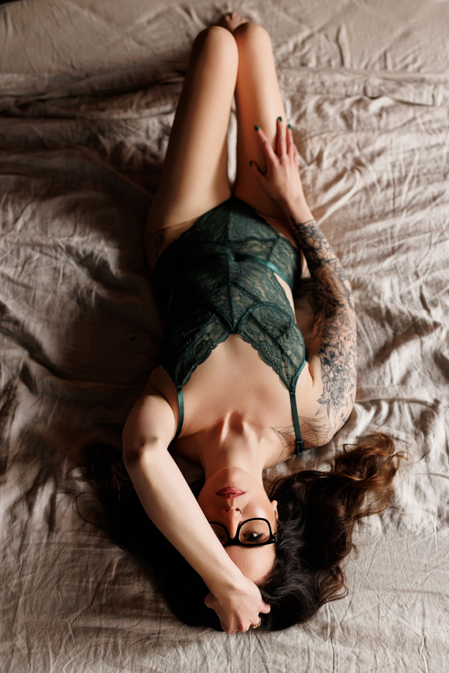 Woman in green lace bodysuit with tattoos lying on bed in artistic boudoir photography style.