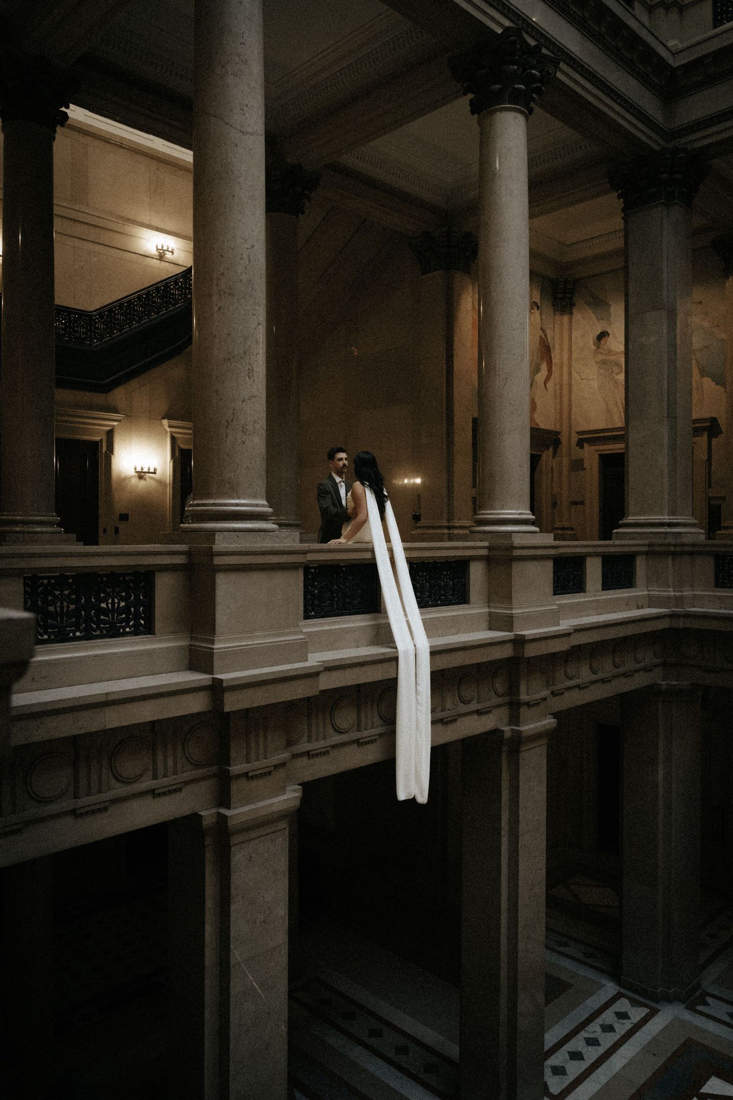 A bride and groom stand on a grand balcony with tall stone columns in an elegant historic building.
