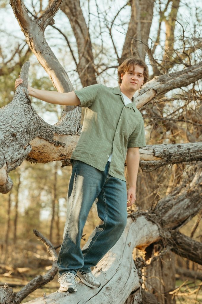 Young man in green shirt and jeans posing on fallen tree branches in a forest setting.
