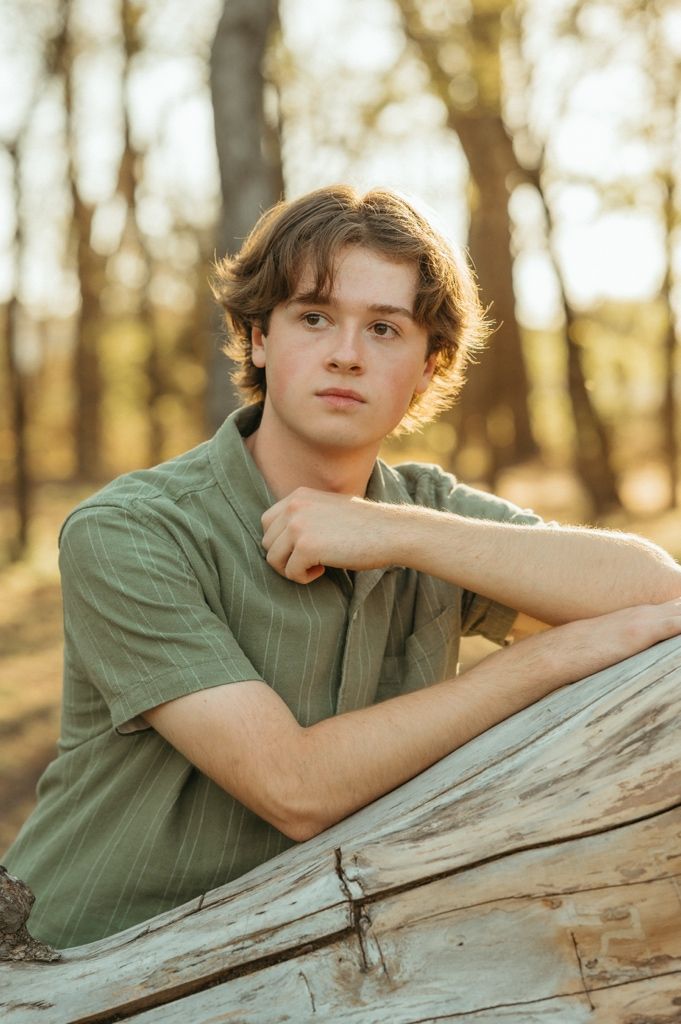 Teen boy in green striped shirt leaning on a wooden log in a sunlit forest setting.