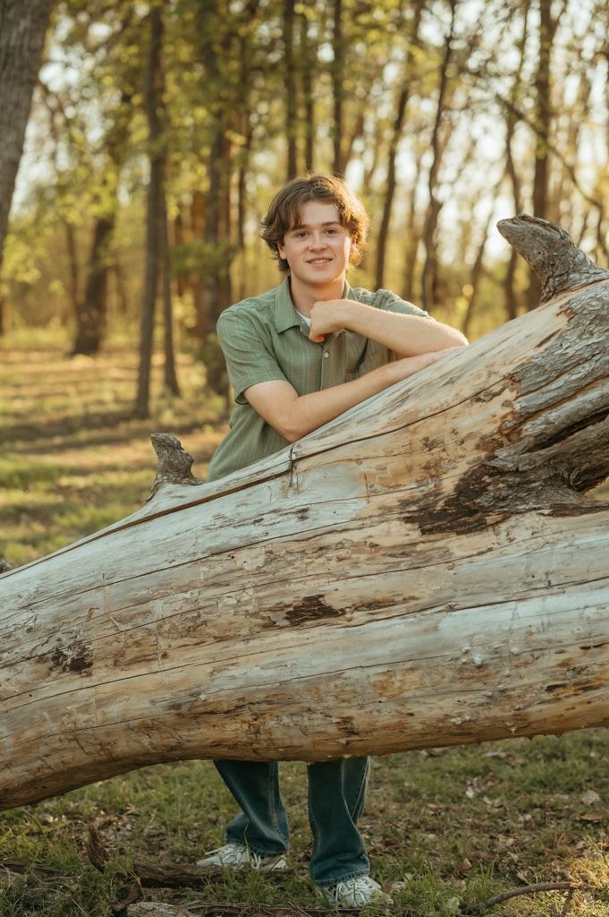 Smiling young man in green polo resting arms on a large fallen log in a forest.