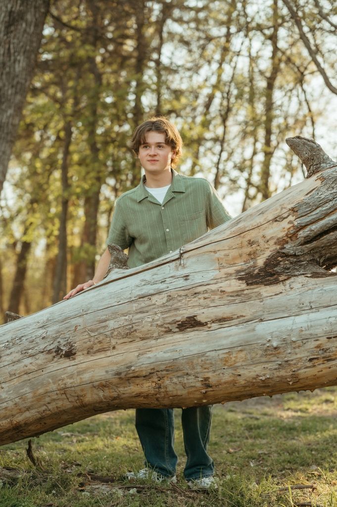 Teen boy in green shirt standing behind a large fallen tree trunk in a wooded area.
