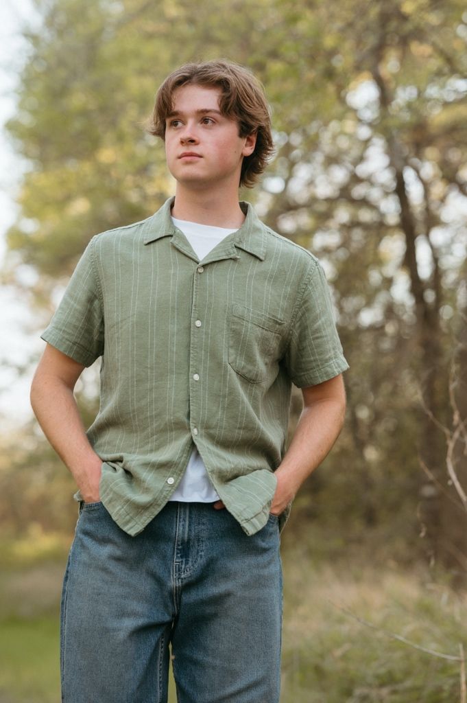Young man in olive green button-up shirt and jeans standing outdoors in a natural wooded area.