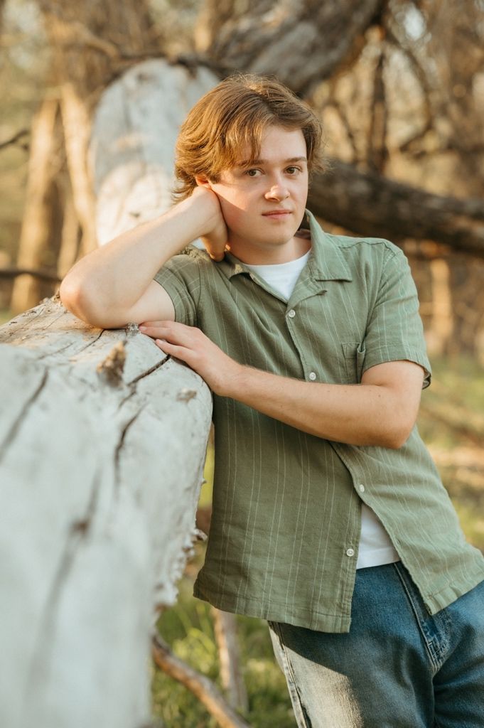Young man with auburn hair leaning on a white fence post outdoors in warm golden light wearing a green shirt.