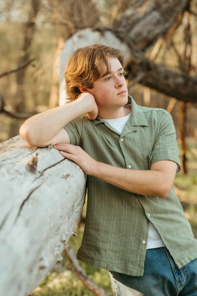 Young man with auburn hair leans on a white log in a sunlit forest, wearing a green button-up shirt.