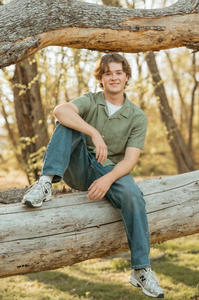 Young man in green shirt and jeans sitting on a log in a wooded outdoor setting, smiling.