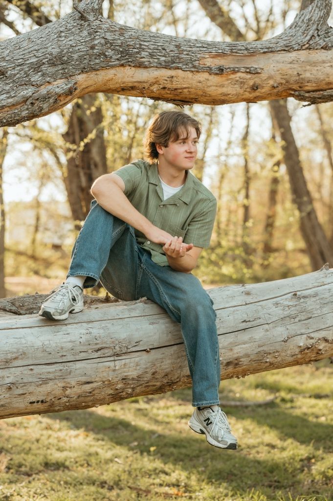 Teen boy in green shirt and jeans sitting on fallen log in autumn forest.