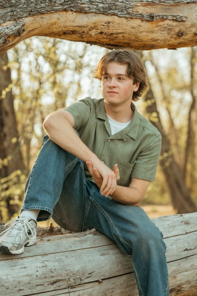 Young man in green shirt and jeans sitting on a fallen log in a sunlit forest.