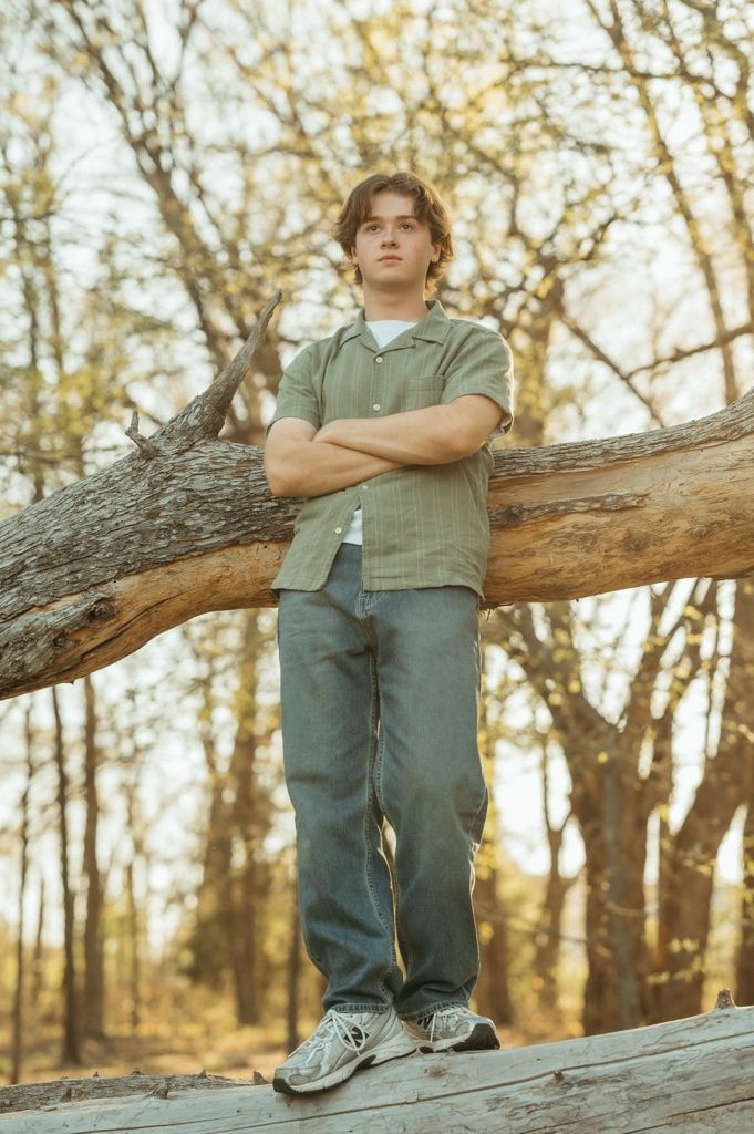 Young man in green shirt with arms crossed leaning against fallen tree outdoors.