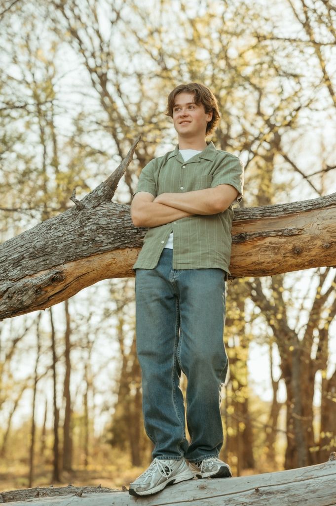 Teen boy in green shirt with arms crossed leaning against a fallen log outdoors.