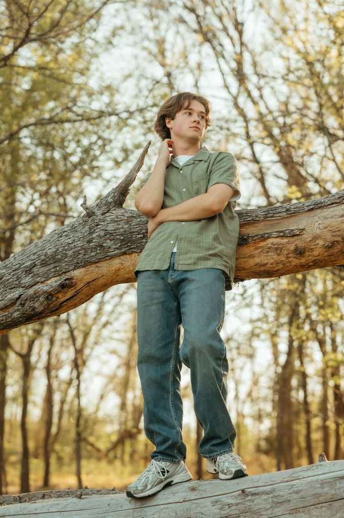 Young man in green shirt and jeans leaning on a fallen log in a forest setting.