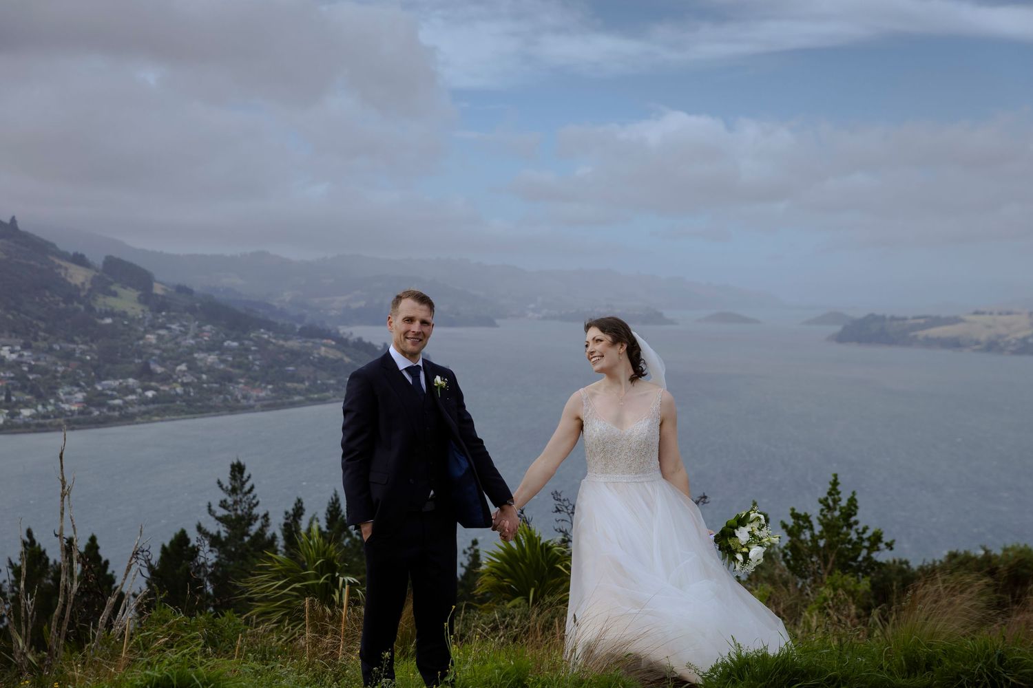 Bride and groom holding hands on a scenic hilltop overlooking a stunning coastal bay on their wedding day.