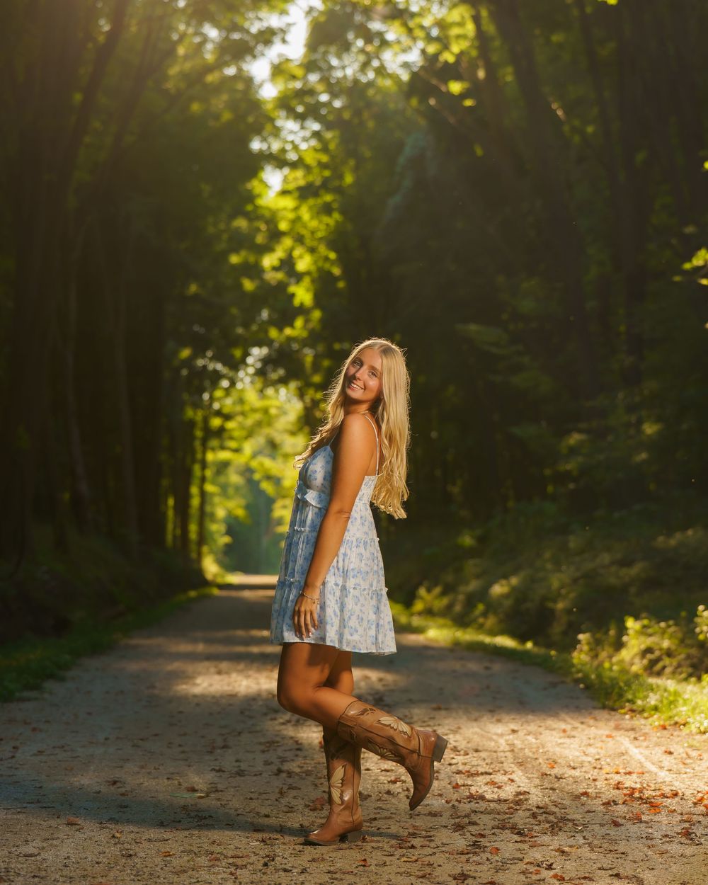 Blonde woman in white dress, denim vest, and cowboy boots posing on a sunlit forest dirt road.