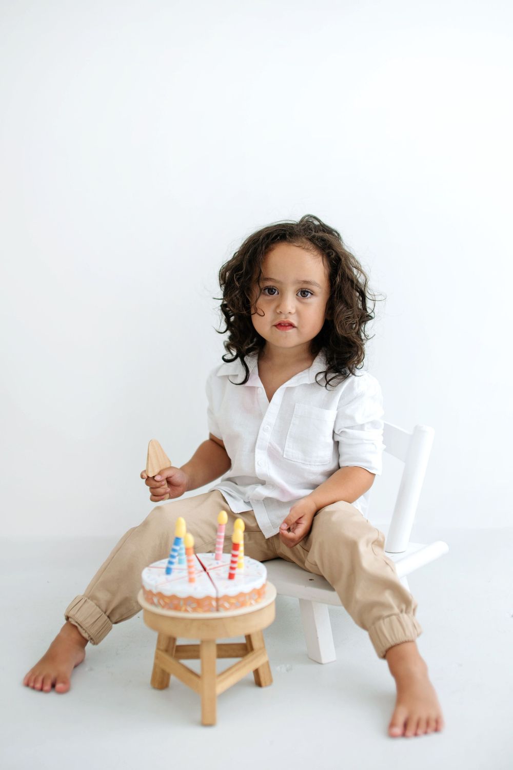 A curly-haired child in white shirt and tan pants sits barefoot beside a small birthday cake with colorful candles on a white background.