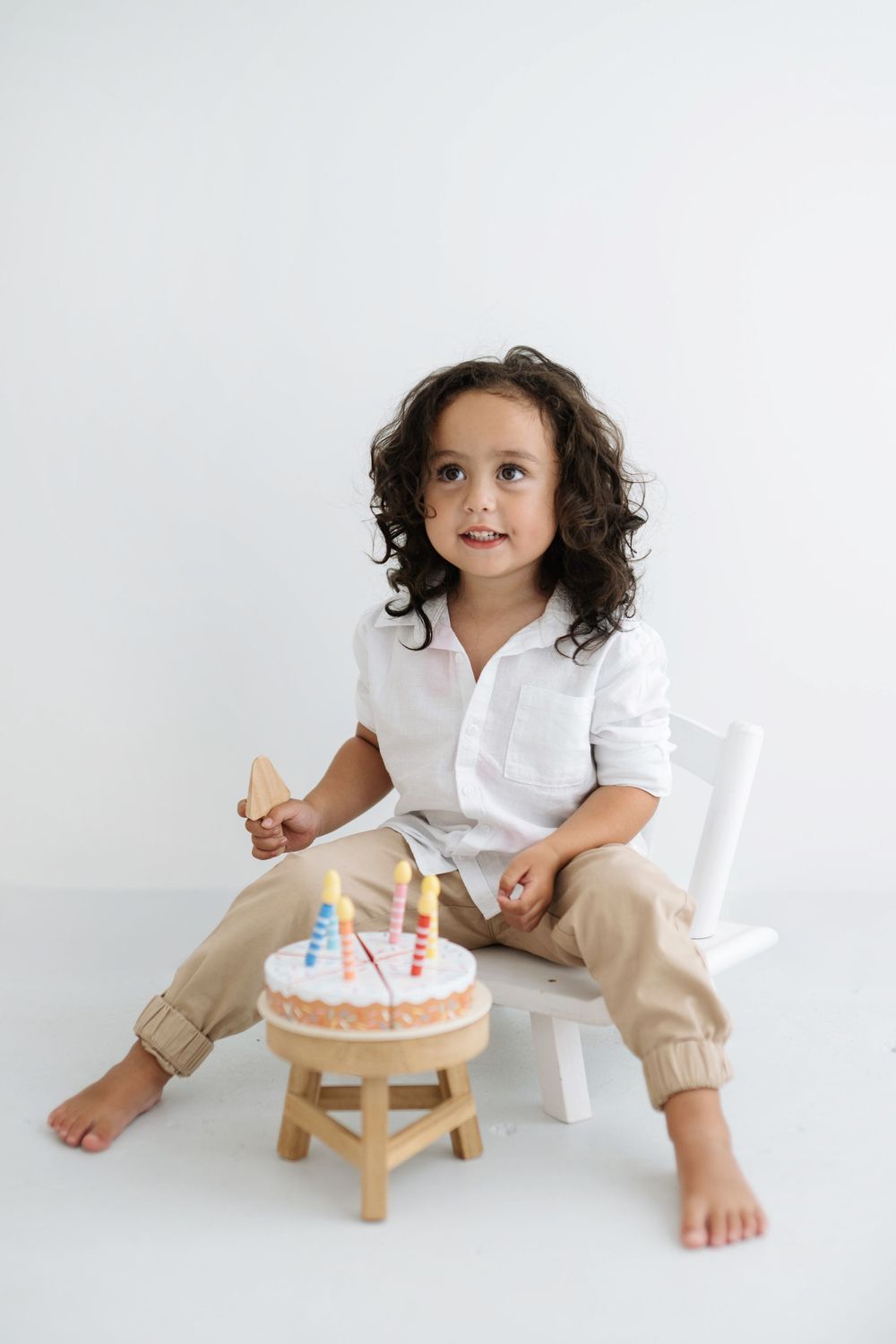 A curly-haired boy in white shirt sits on a chair with a small birthday cake with candles.