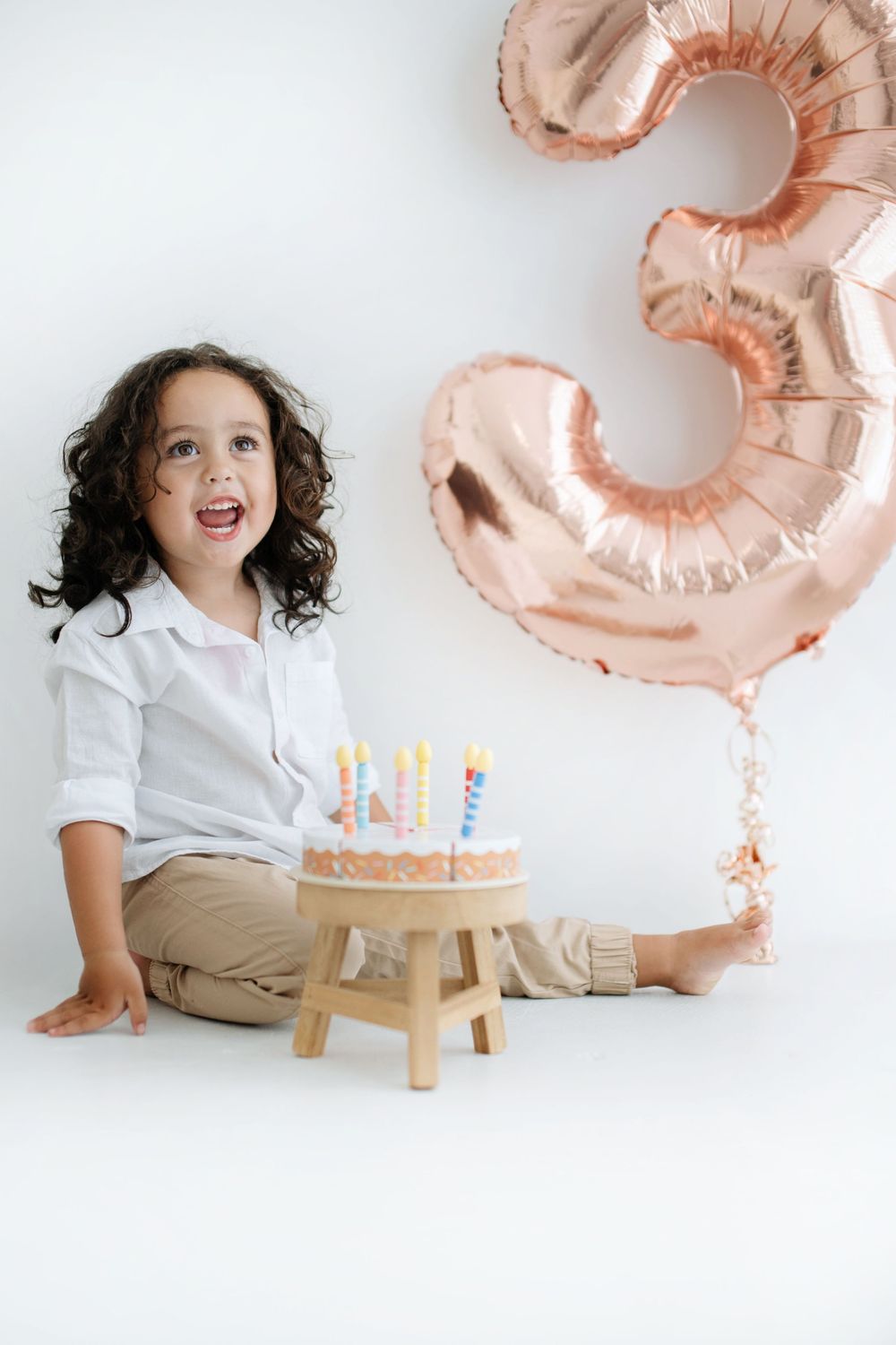 A joyful child celebrates their 3rd birthday with a cake and candles beside a rose gold number 3 balloon.